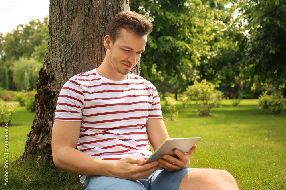 Young man with tablet PC sitting near tree in park