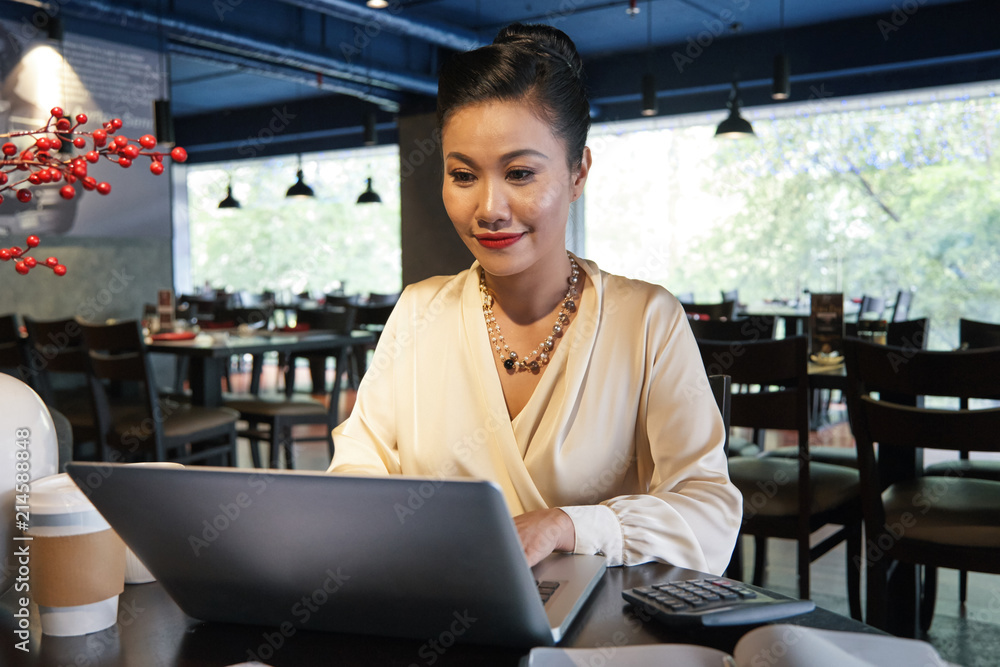 Fototapeta premium Happy Asian businesswoman in white blouse sitting at wooden table in restaurant and using laptop on blurred background 