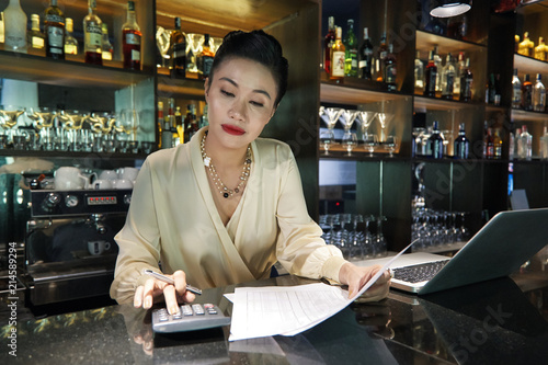 Beautiful Asian woman bartender concentrating on using calculator doing business accounts and holding papers with charts at dark bar counter with laptop placed nearby  photo