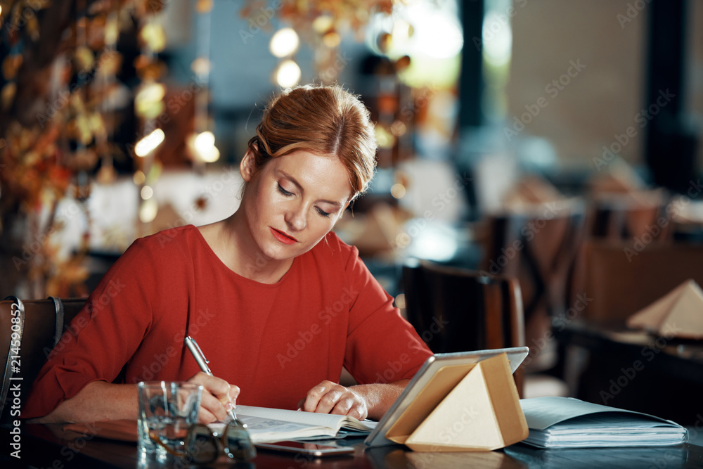 Fototapeta premium Serious female writer working at the cafe while waiting for her order. She is writing ideas in her notebook