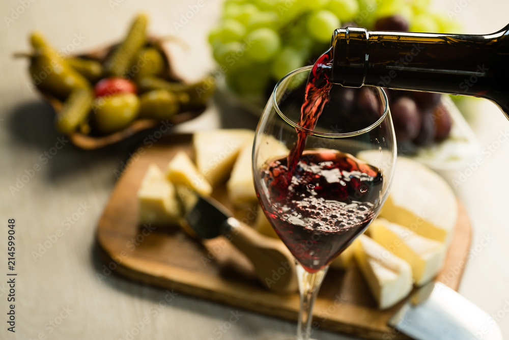 Obraz premium Pouring red wine into the glass in the background a table set with various types of cheese