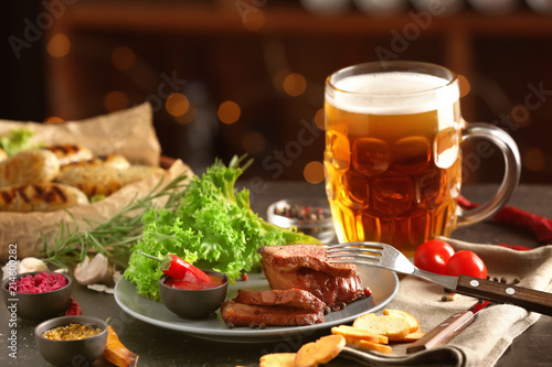 Mug of delicious beer with grilled steak and sauce on table