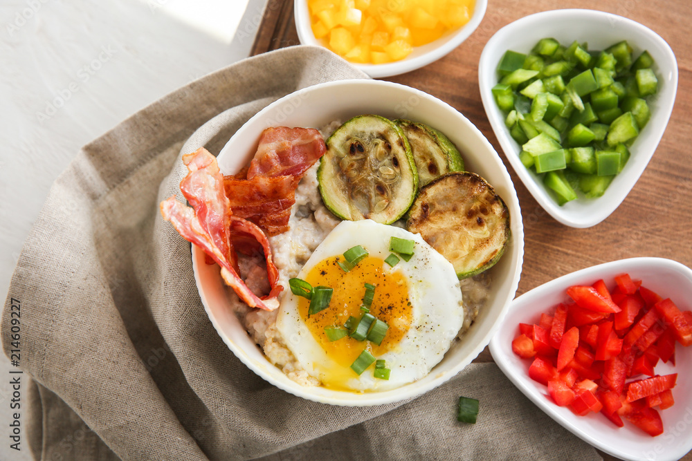 Bowl with tasty oatmeal, fried egg, bacon and vegetables on table