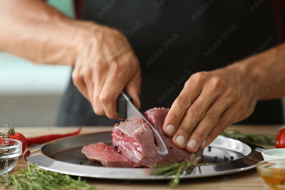 Man cutting raw meat on plate in kitchen, closeup
