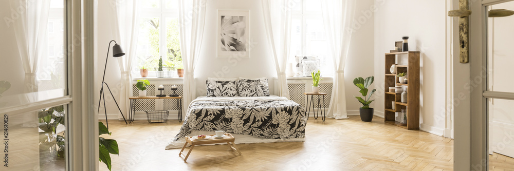 Glass door entrance into a beautiful, bright bedroom interior with breakfast tray on a wooden floor and black floral pattern sheets on a comfy bed - obrazy, fototapety, plakaty 