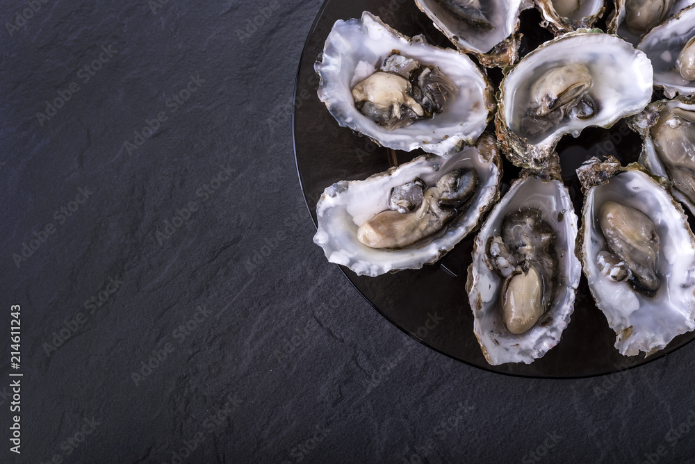Open oysters on black round plate on dark grey background. Fresh Oysters close-up top view. Healthy sea food. Oyster dinner with champagne in restaurant. Copy space Open oysters on black round plate on dark grey background. Fresh Oysters close-up top view. Healthy sea food. Oyster dinner with champagne in restaurant. Copy space