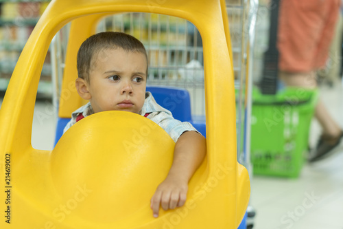 Cute sad little 2 year old baby boy child in the little toy-car trolley at supermarket, Dad or Parent pushing child shopping cart car with his son, Kid first experience concept.