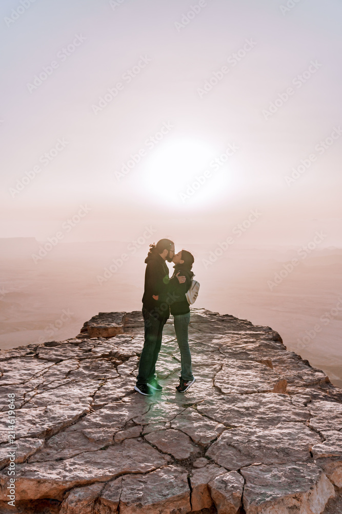 Fototapeta premium Alone young couple of man and women with backpack in israel negev desert admires the view of sunrise. Young pair stands on the edge of the cliff of makhtesh ramon park.
