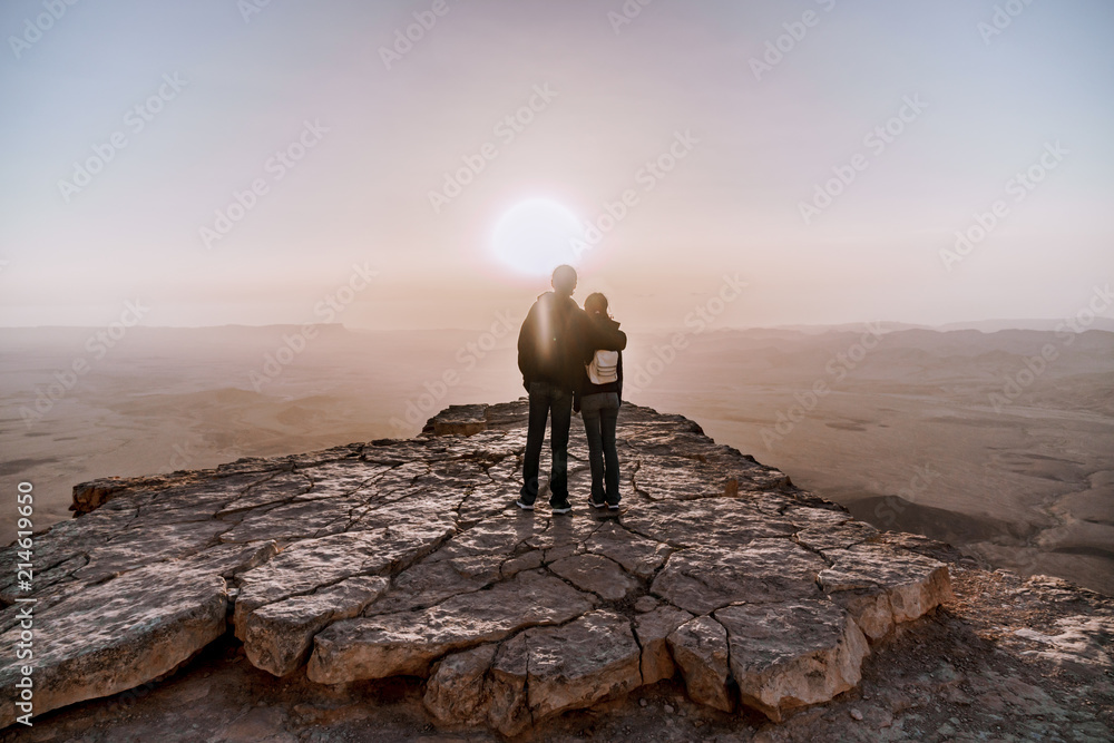 Alone young couple of man and women with backpack in israel negev desert admires the view of sunrise. Young pair stands on the edge of the cliff of makhtesh ramon park. - obrazy, fototapety, plakaty 