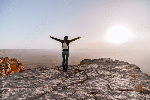 Alone young girl with backpack in israel negev desert admires the view of sunrise. Young female person stands on the edge of the cliff of makhtesh ramon park.