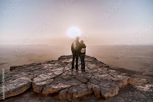 Alone young couple of man and women with backpack in israel negev desert admires the view of sunrise. Young pair stands on the edge of the cliff of makhtesh ramon park. photo