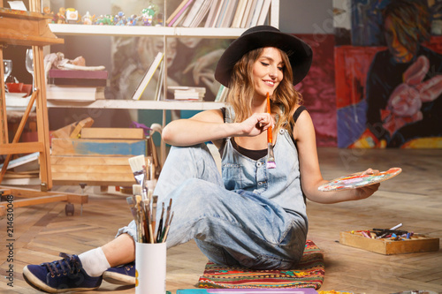 Pretty long-hair female artist in cute black hat looking on palette and holding brush, she sitting on the floor photo