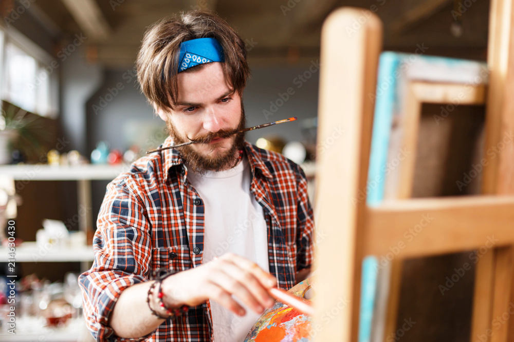 Creative dark-haired bearded hipster male artist in blue bandana holding  palette and drawing behind the easel in his contemporary studio with many  paintings, holding a brush in his teeth Stock Photo
