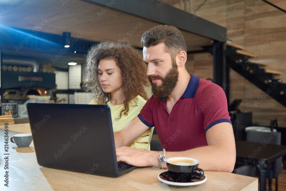 Young man with beard working with a laptop sitting near curly girl in cafe. Drinking coffee, looking concentrated. Boy wearing casual burgundy polo shirt. Students working for educational project.