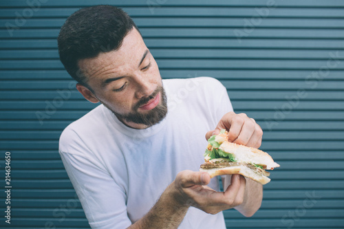 Brunette and bearded man is holding sandwich and showing all vegetables that are in between two pieces of bread. He is looking at it and smiling a bit. Isolated on striped and blue background. photo