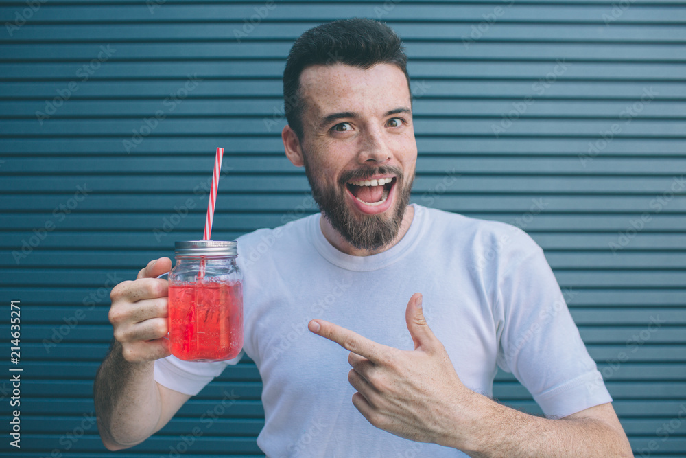 Happy guy is holding glass of lemonade and pointing on it. Also he is smiling and looking on camera. Isolated on striped and blue background.
