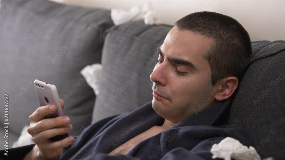 Sick man staying at home and looking at his smartphone while coughing Sick man staying at home and looking at his smartphone while coughing
