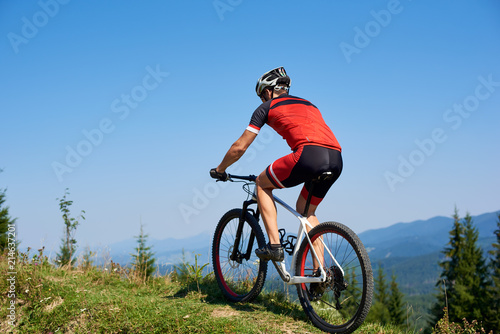 Strong man tourist cyclist in helmet  sunglasses and full equipment riding bike on grassy hill. Mountains and blue summer sky on background. Active lifestyle and extreme sport concept