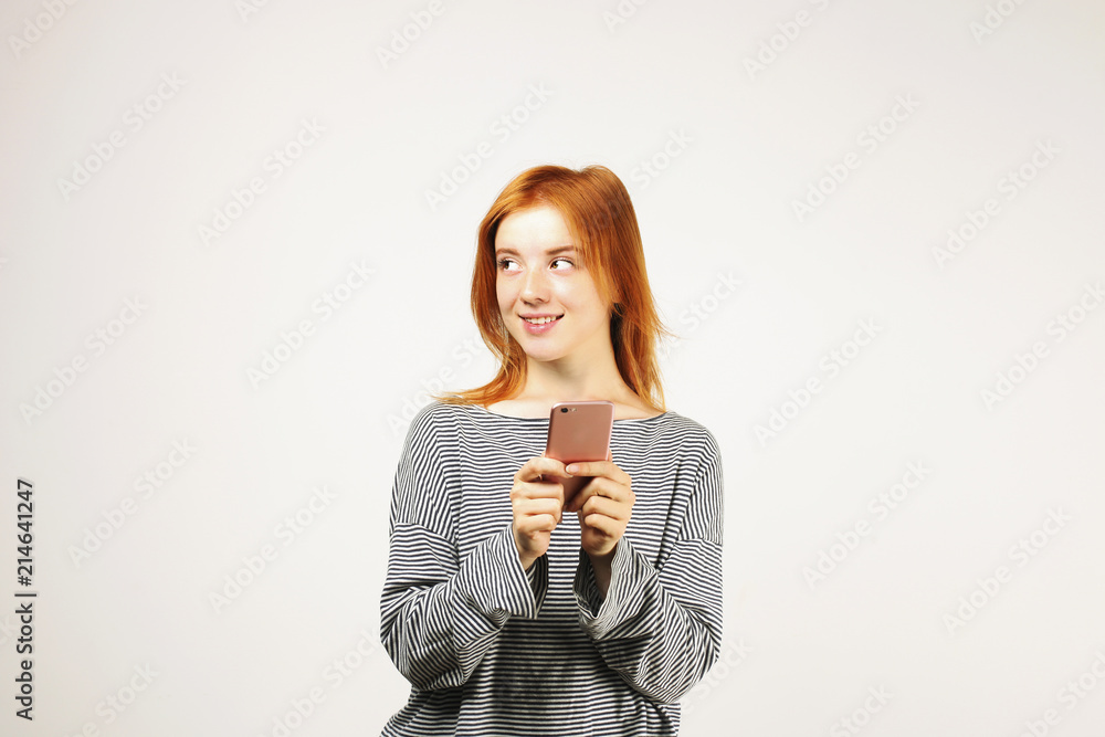 Young attractive natural redhead woman smiling at smartphone screen, using mobile phone, wearing loose gray shirt. White background, copy space. Beautiful female using online dating app concept. Young attractive natural redhead woman smiling at smartphone screen, using mobile phone, wearing loose gray shirt. White background, copy space. Beautiful female using online dating app concept.