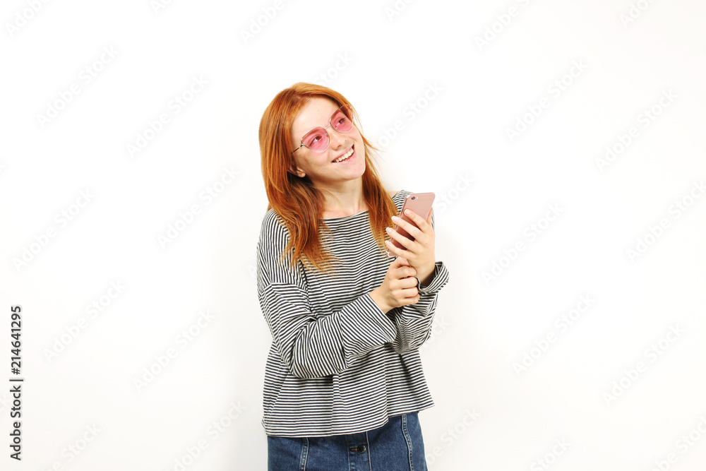 Young attractive natural redhead woman smiling at smartphone screen, using mobile phone, wearing loose grey shirt & cat eye glasses. White copy space background. Female using online dating app concept Young attractive natural redhead woman smiling at smartphone screen, using mobile phone, wearing loose grey shirt & cat eye glasses. White copy space background. Female using online dating app concept