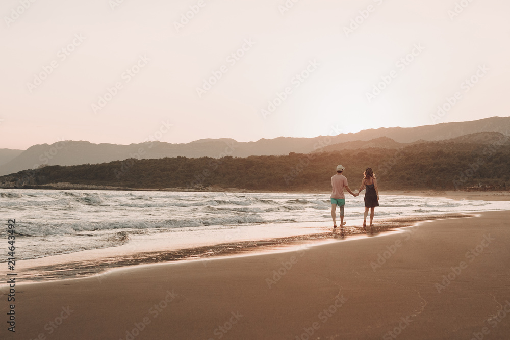 Back view of a romantic hipster couple walking at beach during summer vacation at sunset