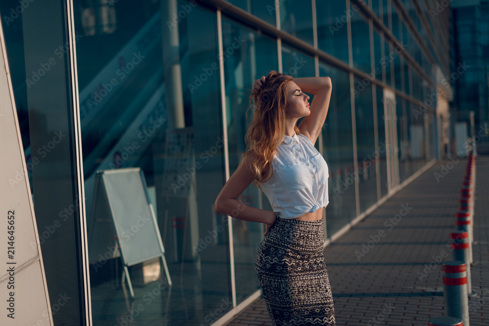 Young attractive blonde woman with curly hair, in stylish outfit posing near the big office center windows. Summer evening