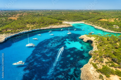 Aerial view of transparent sea with blue water  sandy beach  rocks  green trees  yachts and boats in sunny morning in summer. Travel in Mallorca  Balearic islands  Spain. Top view. Colorful landscape