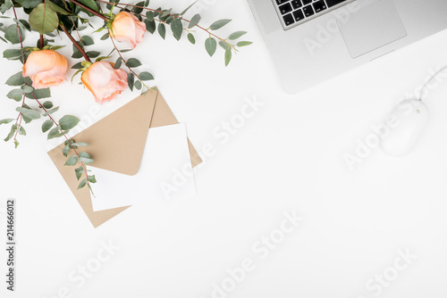Styled feminine desk workspace with pink roses, laptop computer, green eucalyptus leaves, mouse, envelope and white note card. Top view and flat lay of table office desk