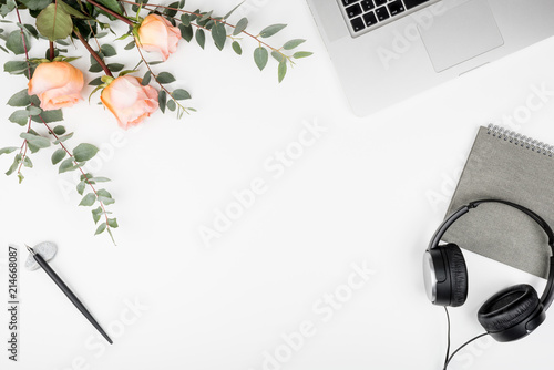 Styled white office desktop with keyboard, earphones, pink roses, eucaliptus branches, notebook and calligraphy pen. Top view of workspace mockup, flat lay, copy space