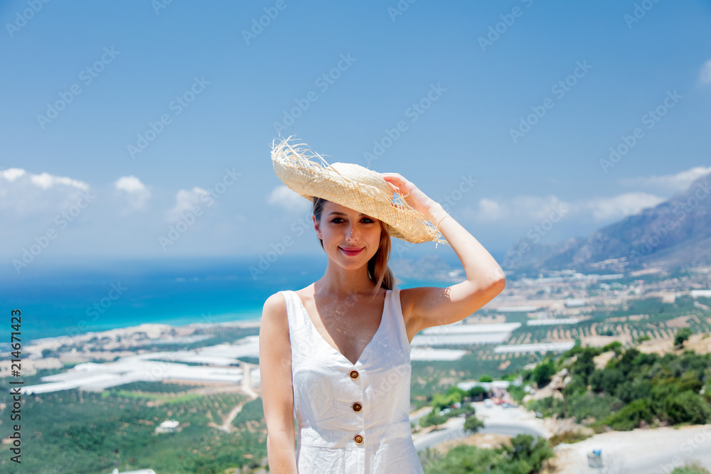 Young woman in white dress and hat is posing on olive garden and sea coast background, Falassandra region, Crete, Greece