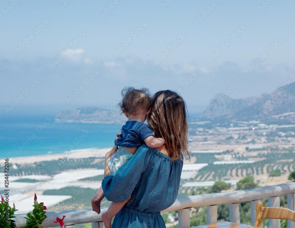 Young caucasian mother with a child have a vacation on Crete, Greece. Mother hold a boy on hands and landscape of a summertime season flowers and olive trees