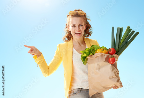 happy woman with paper bag with groceries pointing at something against blue sky