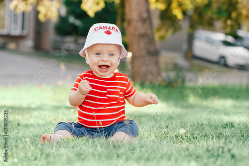 Portrait of little white Caucasian baby boy wearing Canadian hat with maple leaf. Child sitting on grass  in park on July summer  day. Toddler celebrating national Canada day