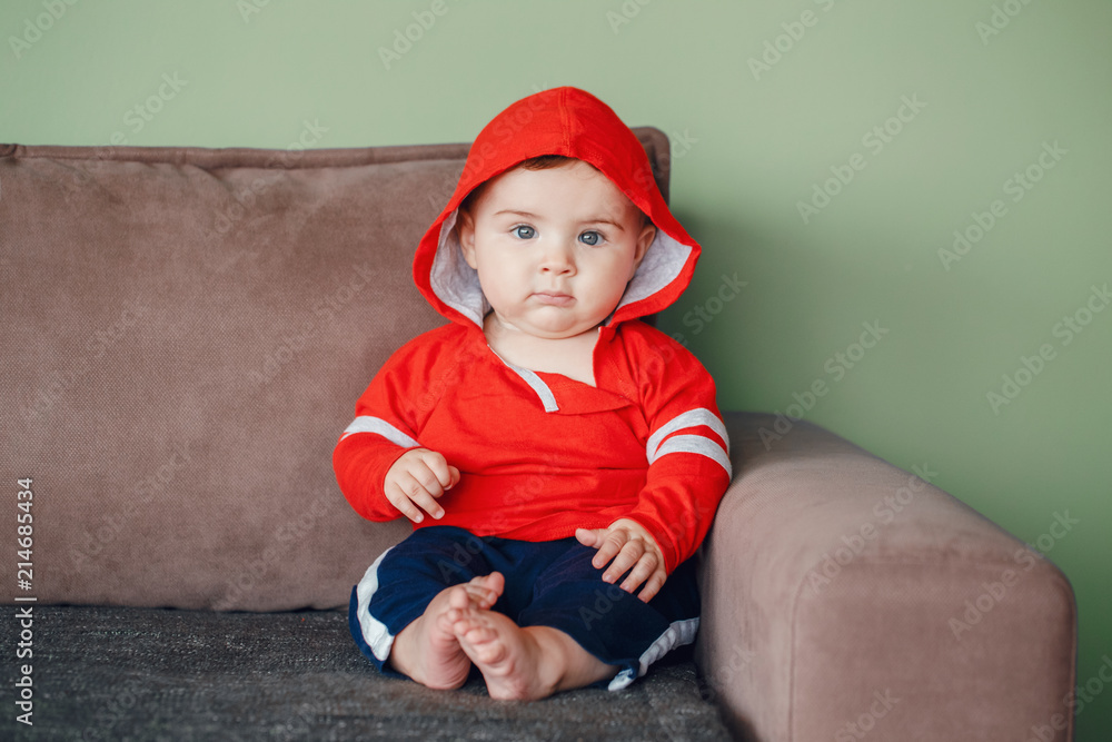 Portrait of cute adorable Caucasian smiling baby boy with blue eyes, wearing red sport hoodie shirt and tracksuit pants. Seven months child sitting on couch at home looking in camera Portrait of cute adorable Caucasian smiling baby boy with blue eyes, wearing red sport hoodie shirt and tracksuit pants. Seven months child sitting on couch at home looking in camera