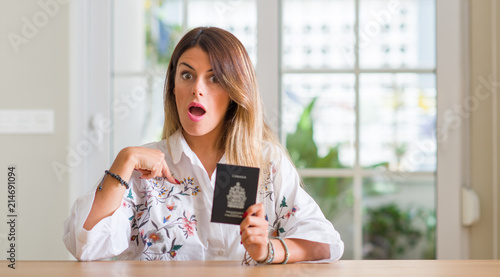 Young woman at home holding a passport of Canada with surprise face pointing finger to himself