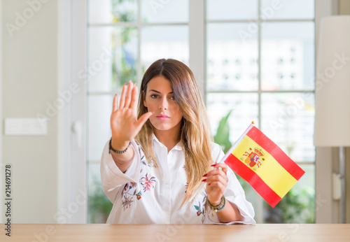 Young woman at home holding flag of Spain with open hand doing stop sign with serious and confident expression, defense gesture