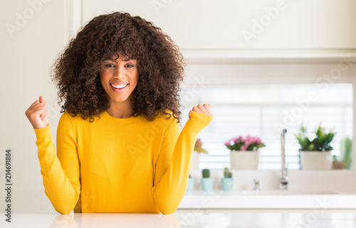 African american woman wearing yellow sweater at kitchen celebrating surprised and amazed for success with arms raised and open eyes. Winner concept.