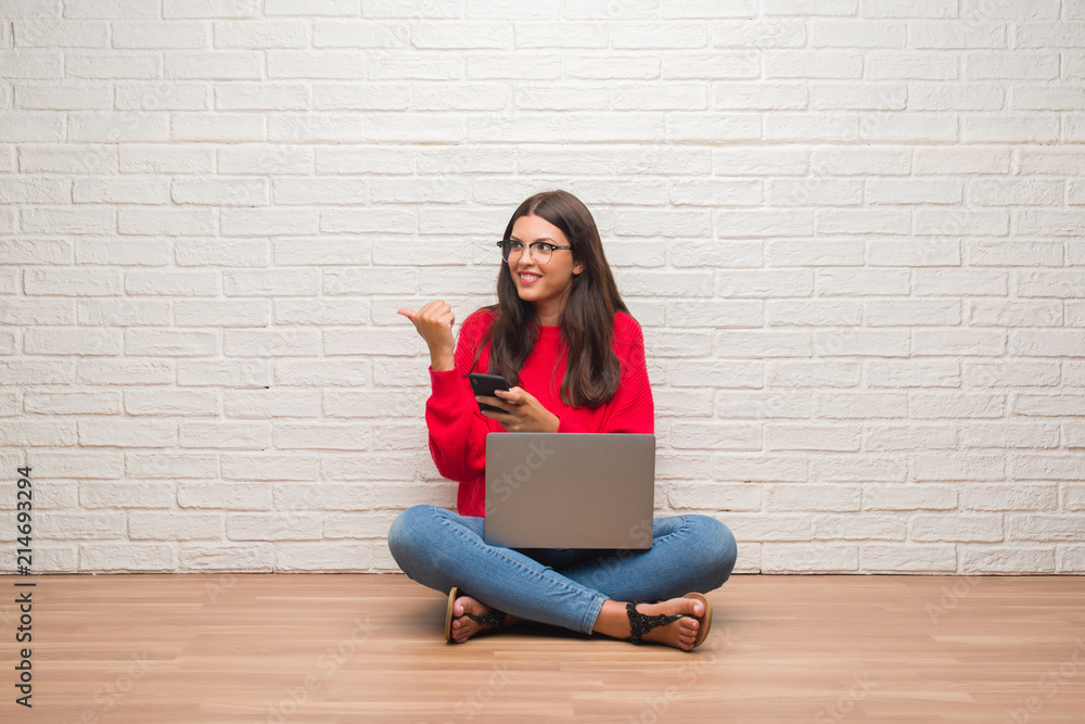 Young brunette woman sitting on the floor using laptop and smartphone pointing and showing with thumb up to the side with happy face smiling Young brunette woman sitting on the floor using laptop and smartphone pointing and showing with thumb up to the side with happy face smiling