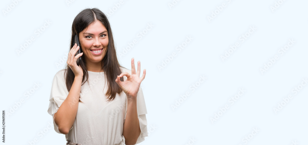 Young beautiful hispanic using smartphone doing ok sign with fingers, excellent symbol Young beautiful hispanic using smartphone doing ok sign with fingers, excellent symbol