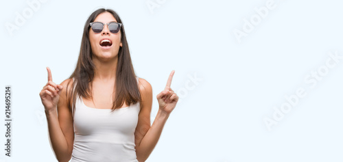 Young beautiful hispanic wearing sunglasses amazed and surprised looking up and pointing with fingers and raised arms.