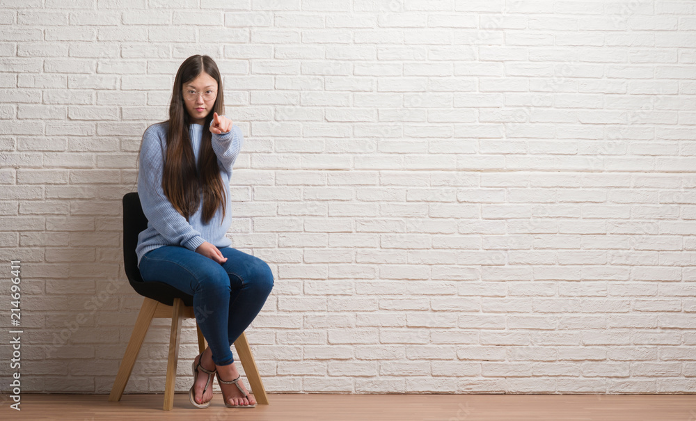 Young Chinese woman sitting on chair over brick wall pointing with finger to the camera and to you, hand sign, positive and confident gesture from the front Young Chinese woman sitting on chair over brick wall pointing with finger to the camera and to you, hand sign, positive and confident gesture from the front