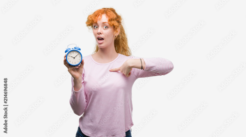 Young redhead woman holding alarm clock with surprise face pointing finger to himself Young redhead woman holding alarm clock with surprise face pointing finger to himself