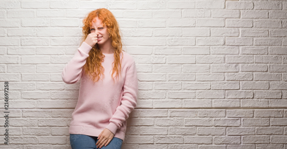 Young redhead woman standing over brick wall smelling something stinky and disgusting, intolerable smell, holding breath with fingers on nose. Bad smells concept. Young redhead woman standing over brick wall smelling something stinky and disgusting, intolerable smell, holding breath with fingers on nose. Bad smells concept.