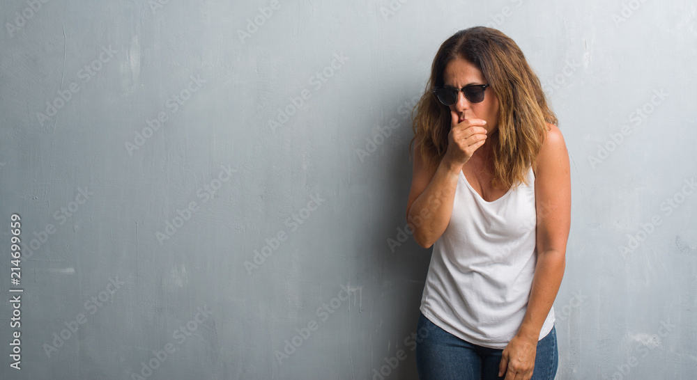 Middle age hispanic woman over grey wall wearing sunglasses feeling unwell and coughing as symptom for cold or bronchitis. Healthcare concept.