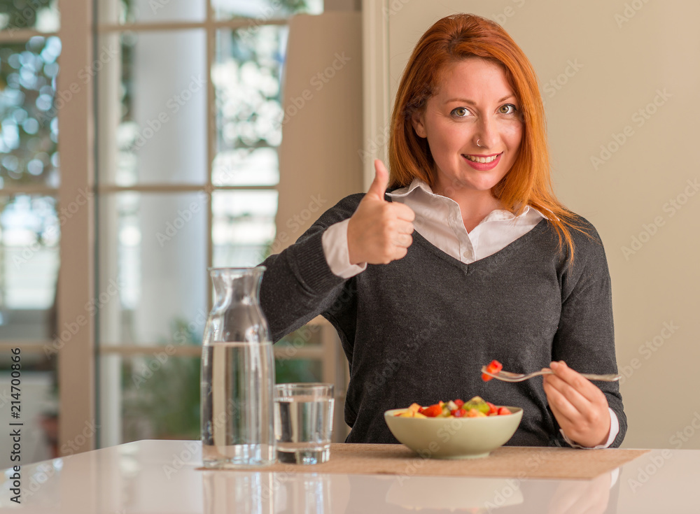 Redhead woman eating fruit bowl, kiwi and strawberry at home happy with big smile doing ok sign, thumb up with fingers, excellent sign Redhead woman eating fruit bowl, kiwi and strawberry at home happy with big smile doing ok sign, thumb up with fingers, excellent sign