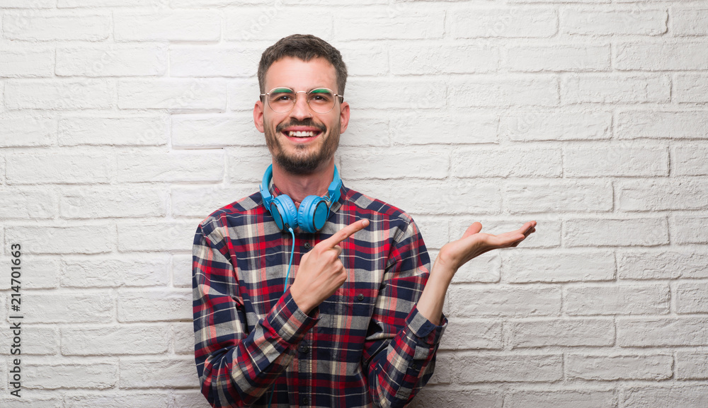Young adult man over brick wall wearing headphones very happy pointing with hand and finger Young adult man over brick wall wearing headphones very happy pointing with hand and finger