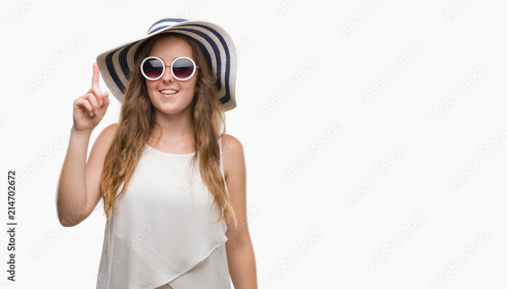 Young blonde woman wearing sunglasses and summer hat surprised with an idea or question pointing finger with happy face, number one Young blonde woman wearing sunglasses and summer hat surprised with an idea or question pointing finger with happy face, number one