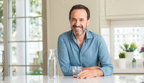 Middle age man drinking a glass of water with a happy face standing and smiling with a confident smile showing teeth photo
