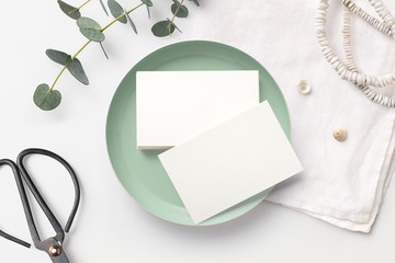 Minimalist feminine branding / business card mockup with stack of cards in a small bowl, scissors, boho necklace and eucalyptus twigs on a styled desk. Mint, white and neutral hues. Flat lay.