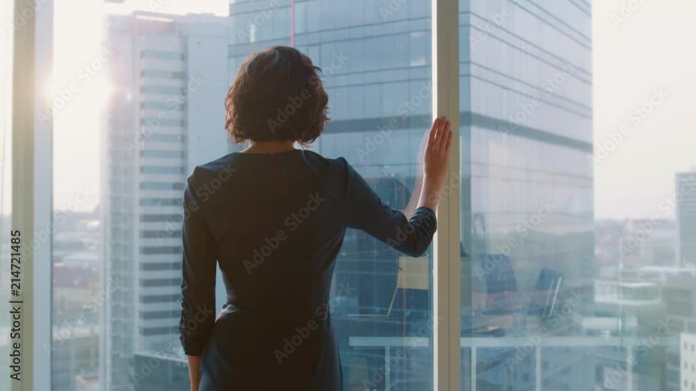 Back View Shot of Successful Businesswoman in the Striking Black Dress Standing in Her Office and Looking out of the Window Thoughtfully. Back View Shot of Successful Businesswoman in the Striking Black Dress Standing in Her Office and Looking out of the Window Thoughtfully.